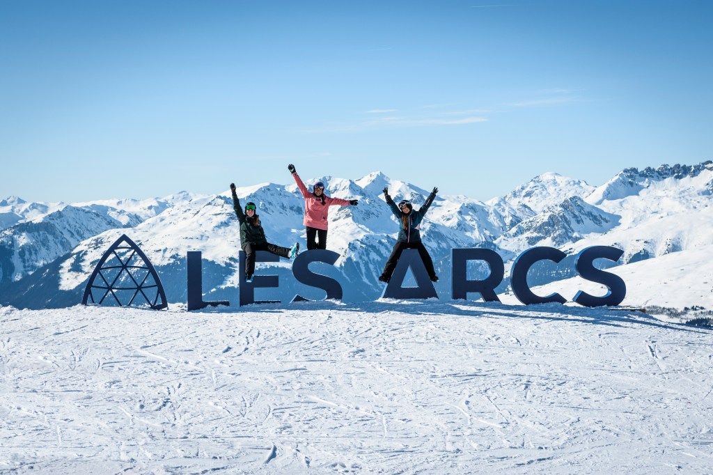 Three people dressed in ski gear are jumping in front of big letters which say 'Les Arcs'. The background is snowy mountains of Paradiski. Photo credit: Juliette Rebour